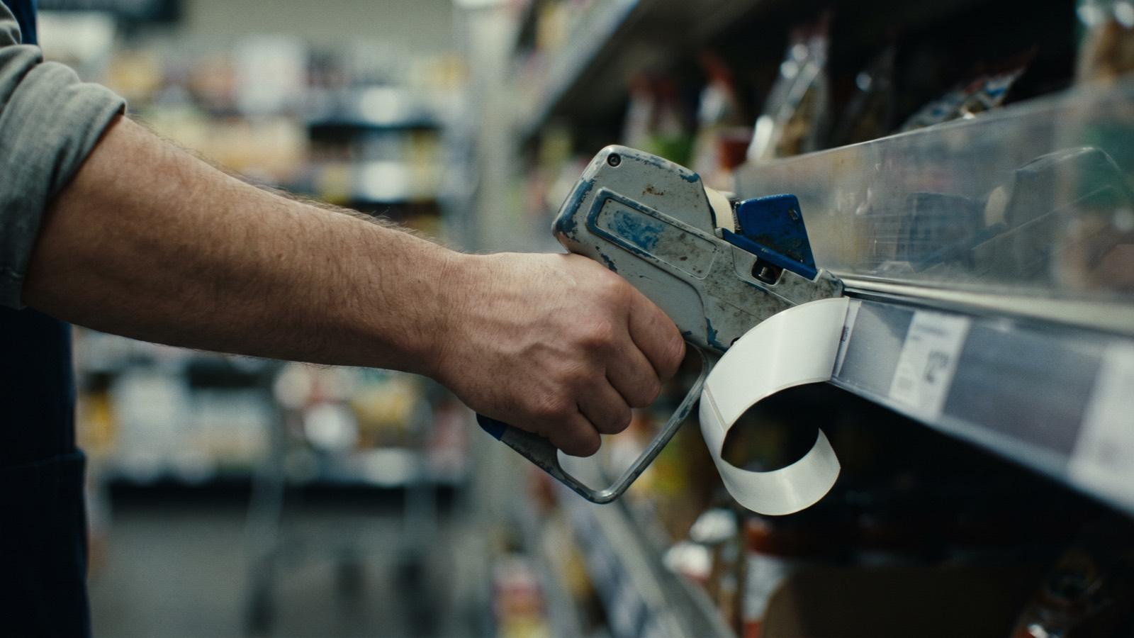 A hand holding a label gun against a shelf edge, a blank price label curling off the roll